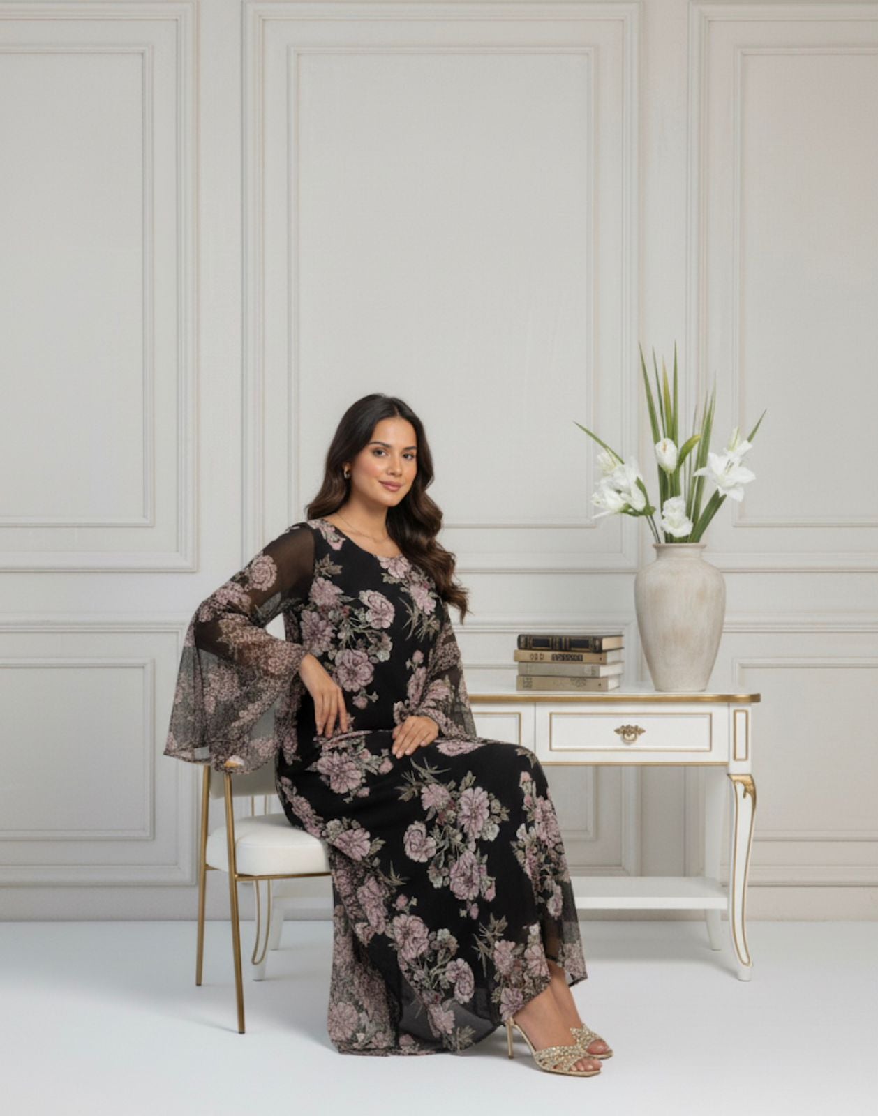 Woman in a floral dress sitting in a stylish room with a vase of flowers on a table.