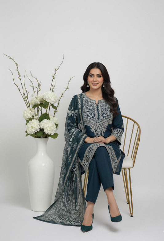 Woman in a blue embroidered outfit sitting on a chair with a vase of flowers next to her.