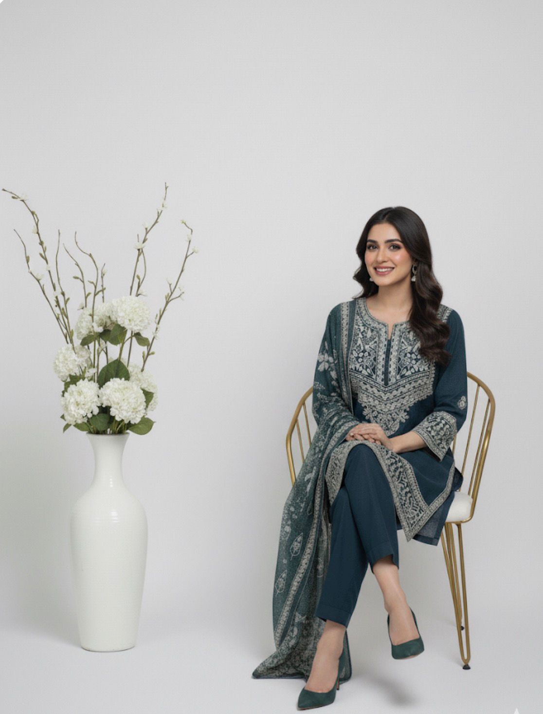 Woman in a blue and green patterned outfit sitting on a chair with a vase of flowers next to her.